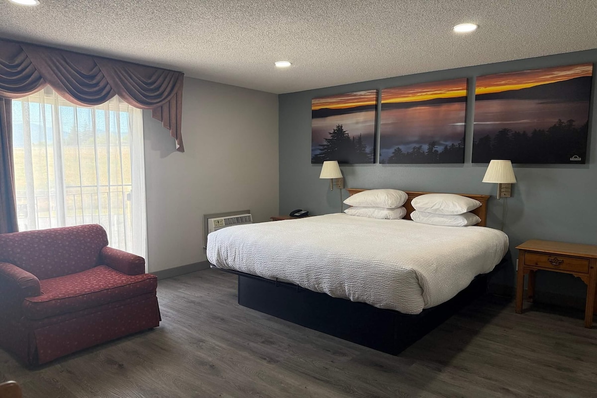 A bedroom with a bed, a chair, a bedside table, a lamp, and a painting on the wall.