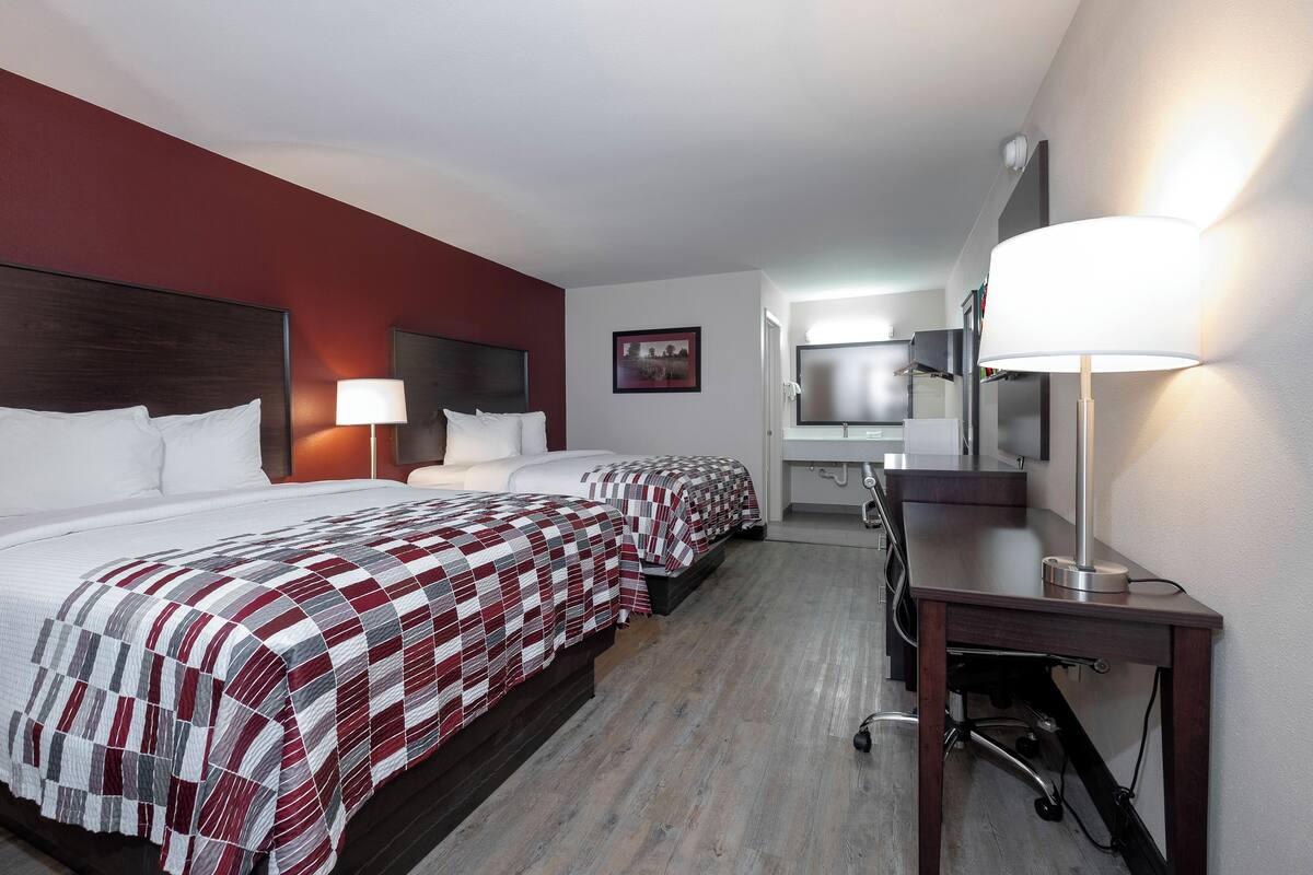 A hotel room with two beds, a desk, a chair, a lamp, a TV, and a bathroom.