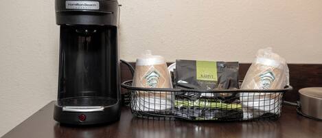 A coffee maker and a basket containing coffee pods and a branded coffee mug on a wooden table.