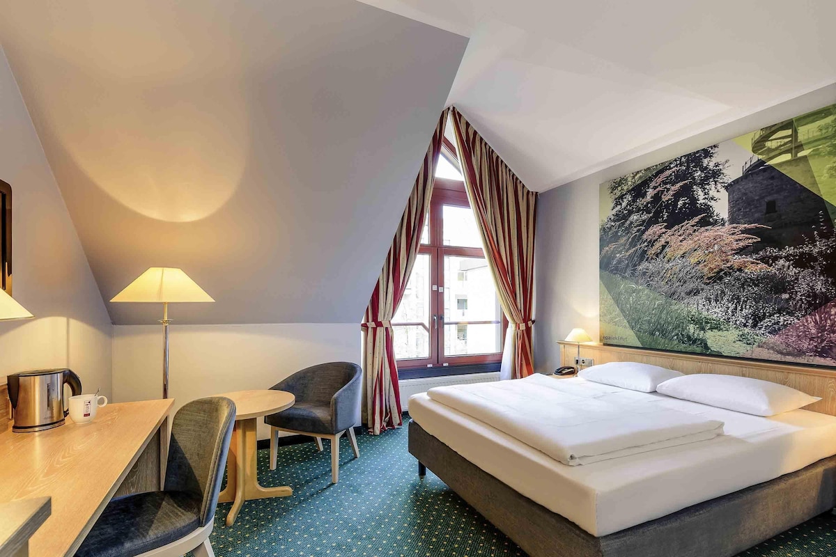 Une chambre d’hôtel avec un lit, un bureau et une chaise, une lampe et une grande fenêtre avec des rideaux.