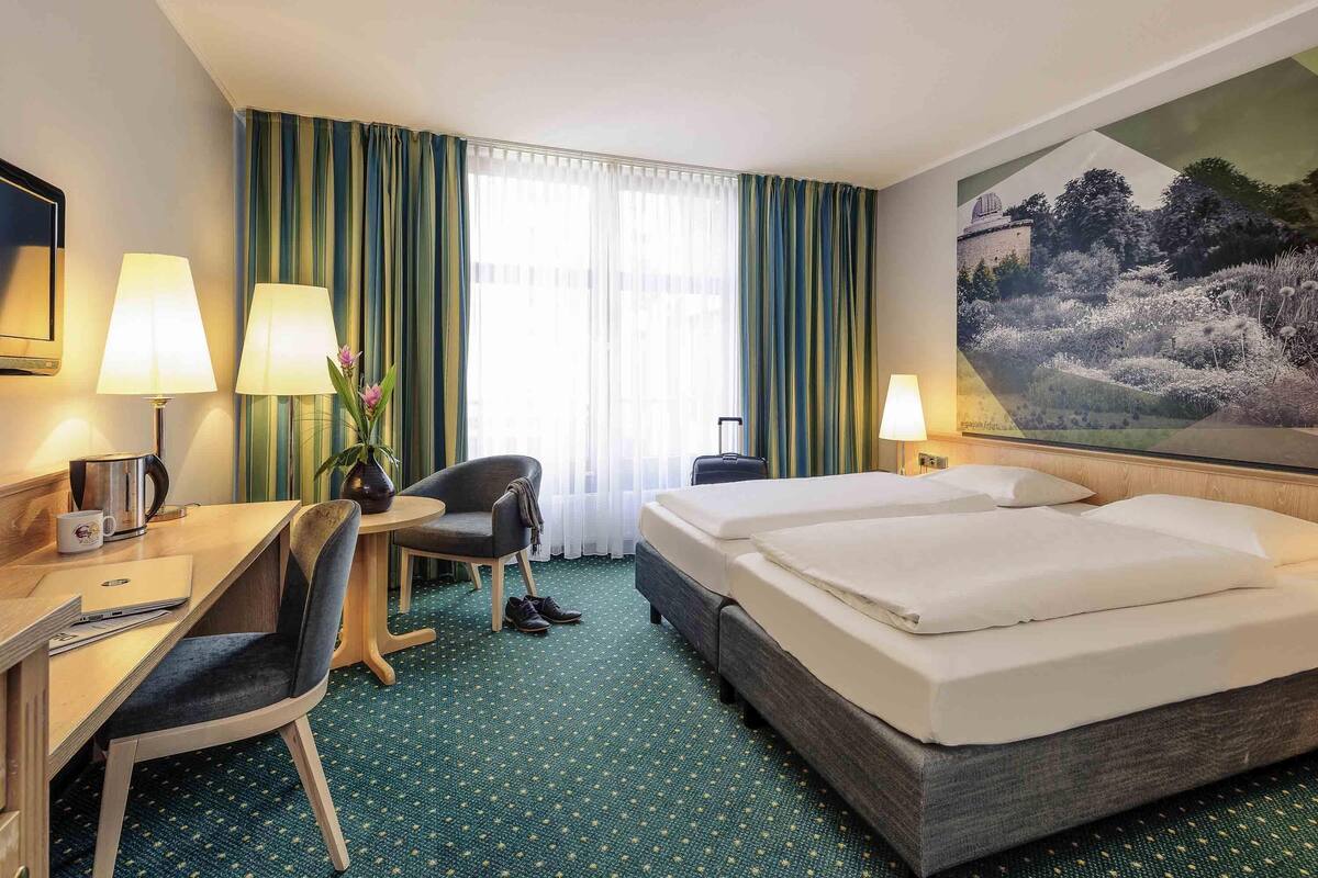 Habitación de hotel con cama, escritorio, silla, lámparas y un cuadro en la pared.