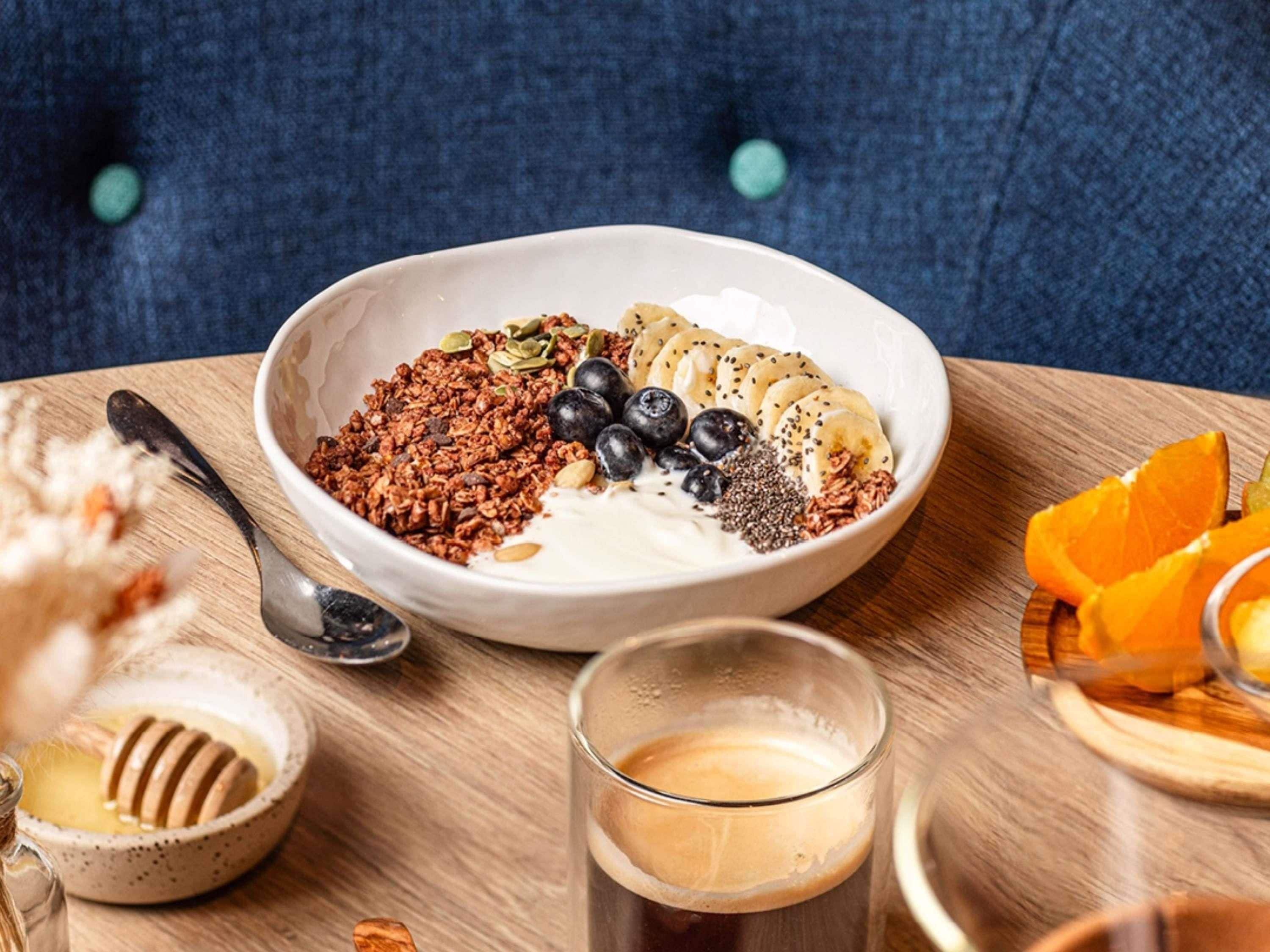 Un bol de granola accompagné de yaourt, de myrtilles, de rondelles de banane et de graines, posé sur une table en bois.