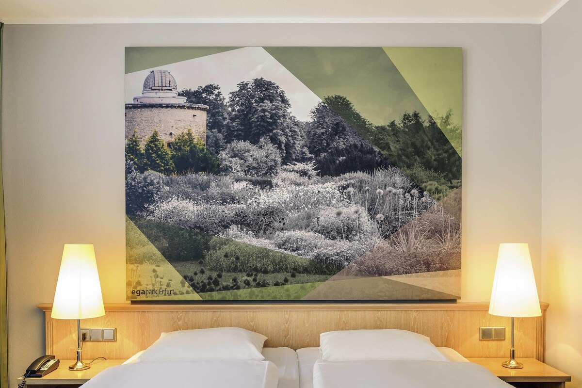 Une chambre d’hôtel avec deux lits, une table de chevet avec une lampe, un téléphone et un grand tableau représentant un paysage accroché au mur.
