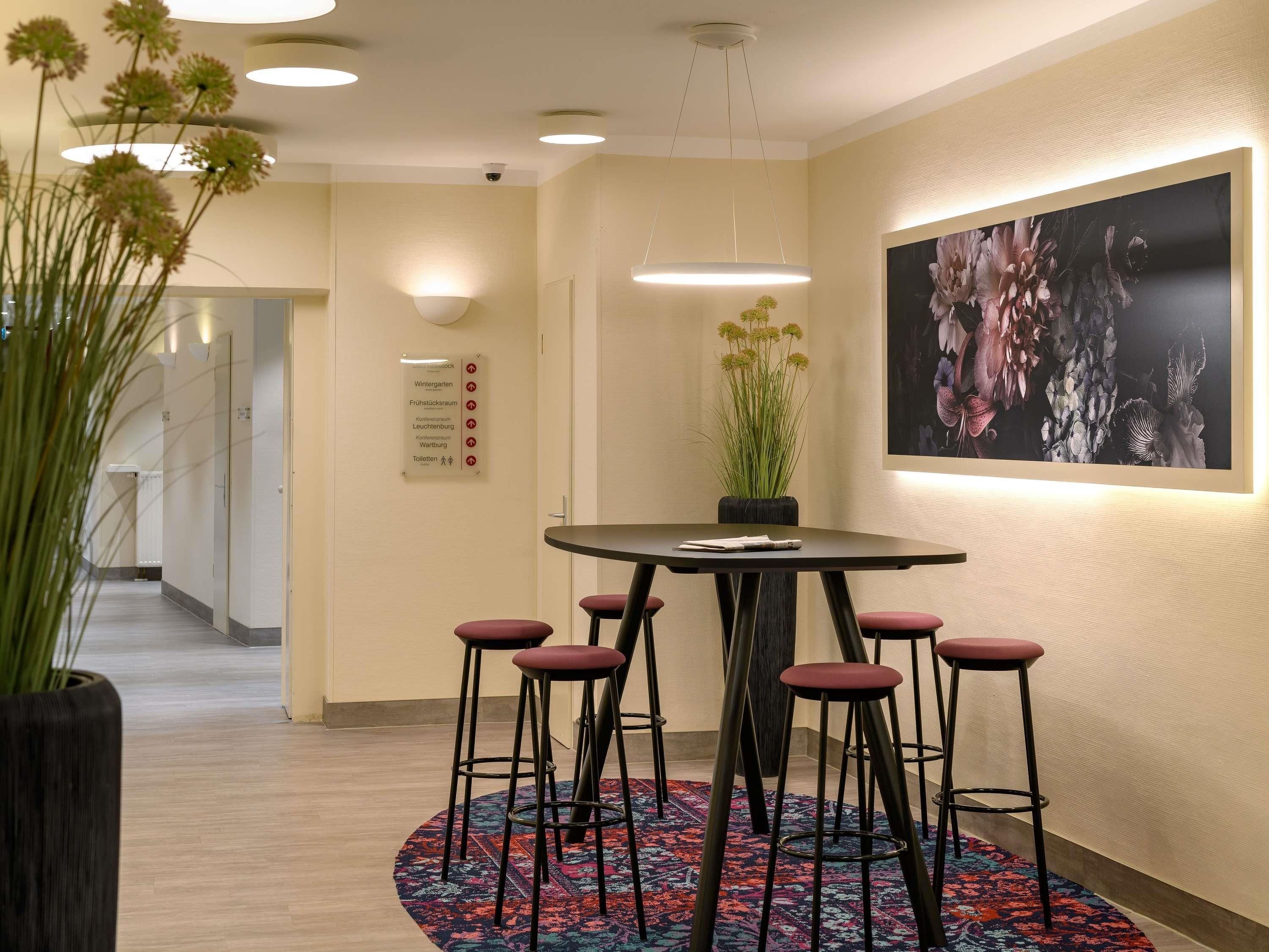 A hotel lobby with a round table, bar stools, a potted plant, a directional sign, and a large floral artwork on the wall.