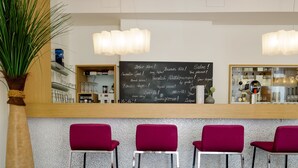 Una zona de bar con una pizarra mostrando saludos en varios idiomas, tres taburetes rojos y una planta en maceta.