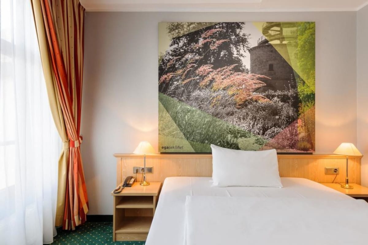 Een hotelkamer met een bed, twee nachtkastjes, een telefoon en een landschapsschilderij aan de muur.