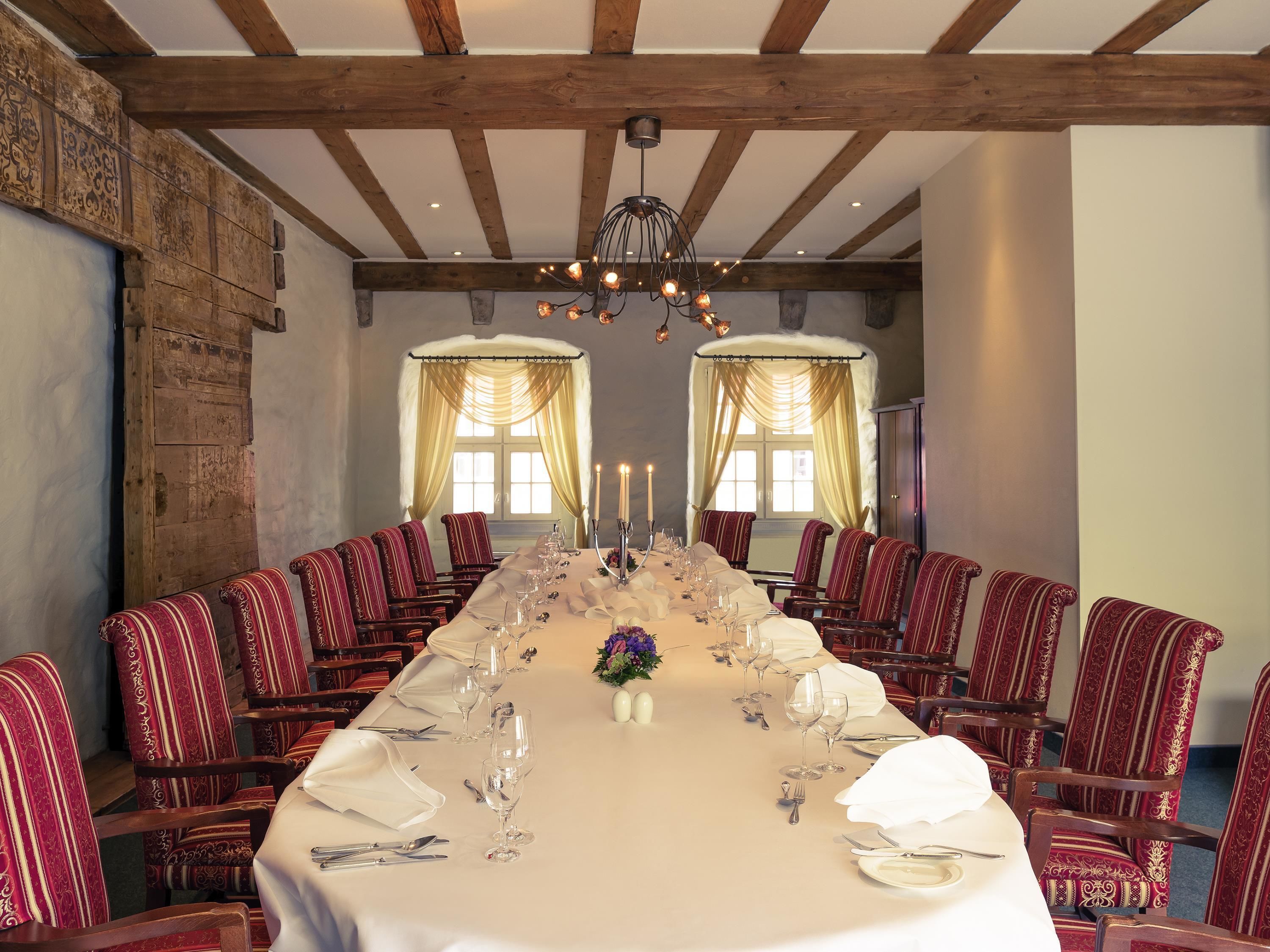A long dining table set for a formal meal, flanked by red upholstered chairs, with a chandelier above and windows with curtains on either side.