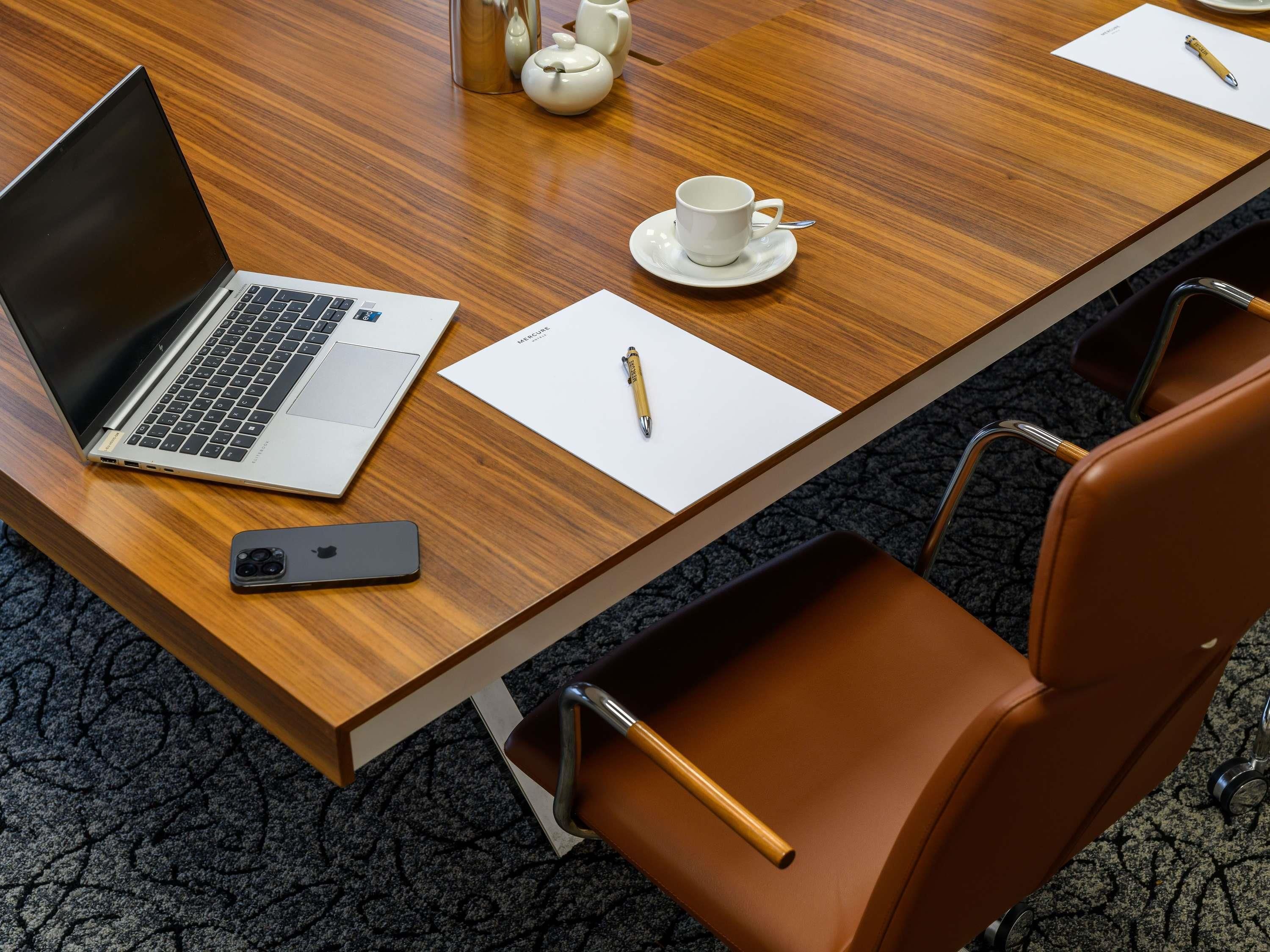 Une table de conférence en bois sur laquelle sont posés un ordinateur portable, un téléphone intelligent, une théière, une tasse et un stylo, le tout sur une feuille de papier blanche.