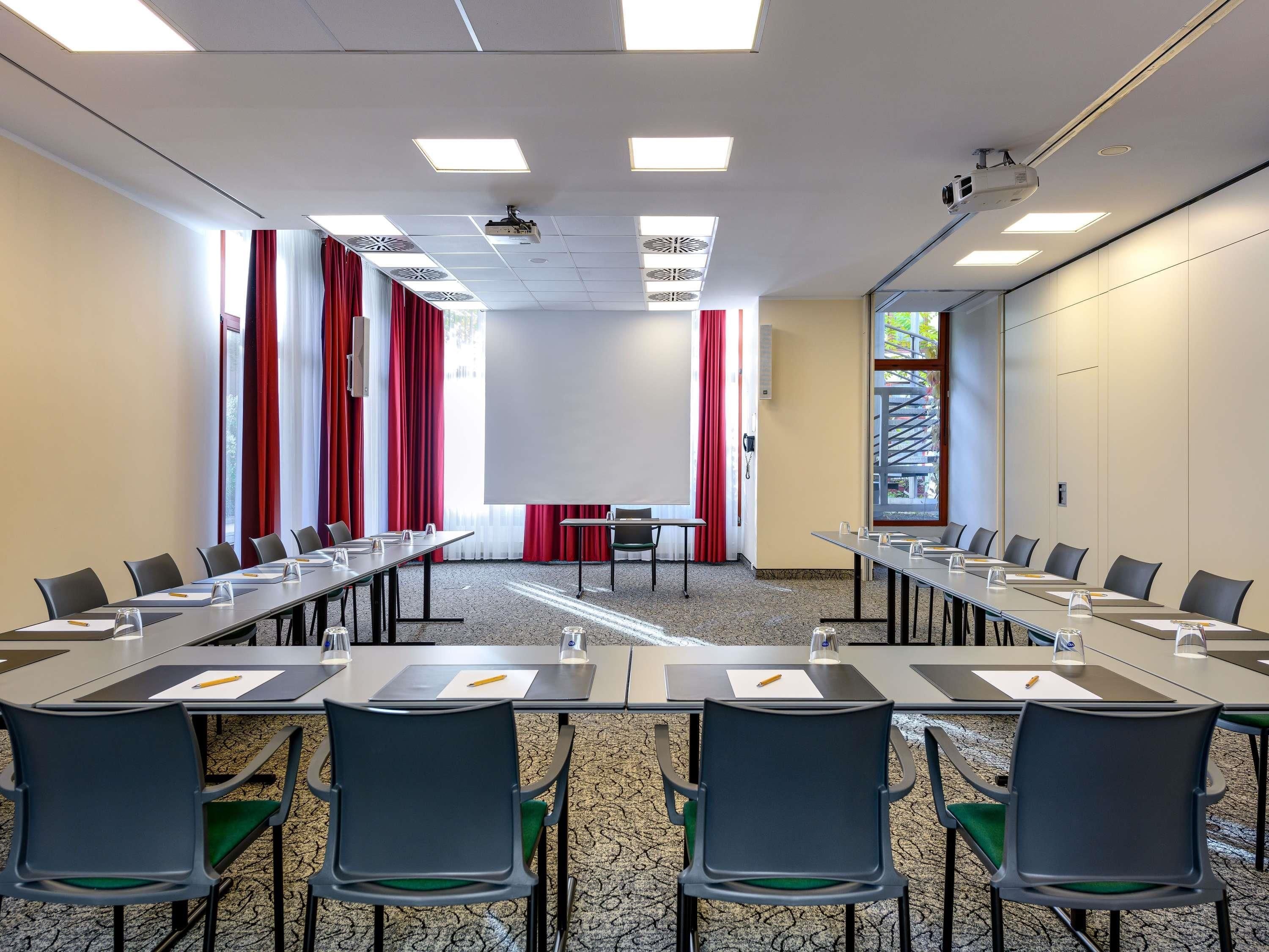 Ein Konferenzraum mit langen Tischen, Stühlen, einem Projektor und einer großen Leinwand.