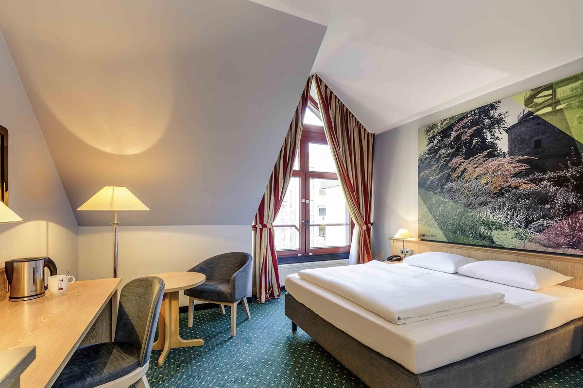 Une chambre d’hôtel avec un lit, un bureau avec une chaise, une lampe et une grande fenêtre avec des rideaux.