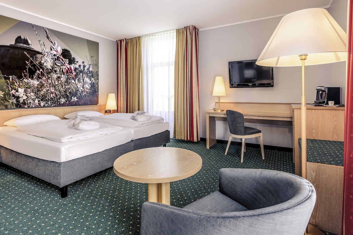 A hotel room with a bed, desk, chair, TV, and artwork.