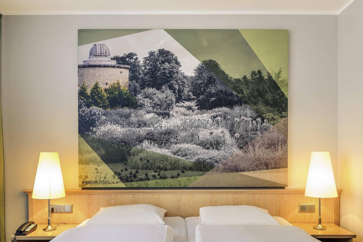 Habitación de hotel con dos camas, una mesita de noche con lámpara, teléfono y un cuadro de paisaje grande en la pared.