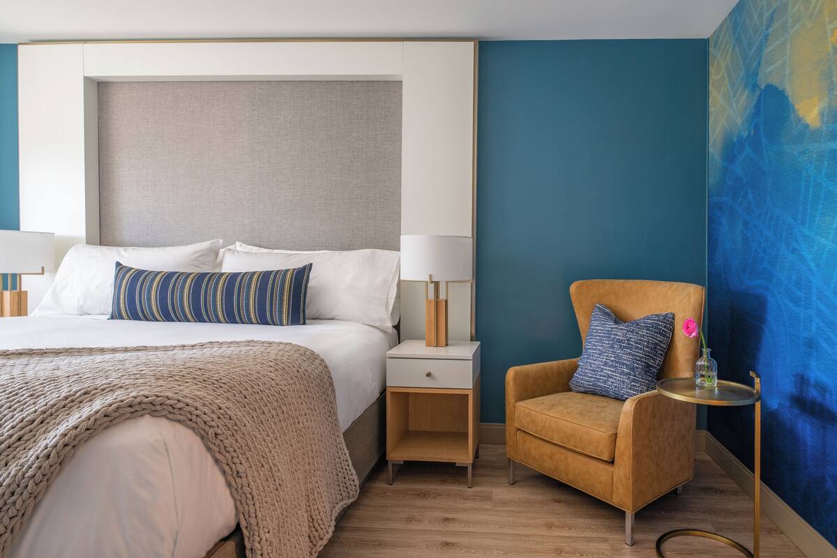 A hotel room with a bed, bedside table, chair, and a large abstract painting on the wall.
