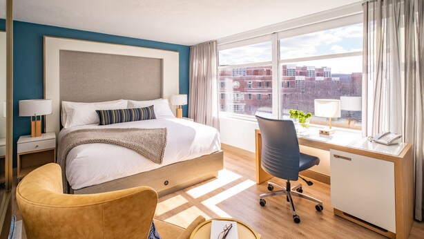 A hotel room with a bed, a desk, a chair, and a view of buildings through large windows.