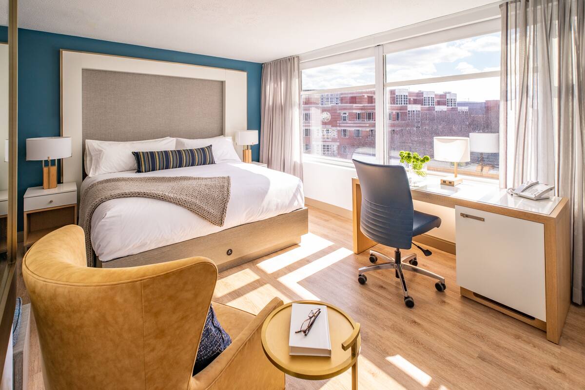 A hotel room with a bed, a desk, a chair, and a view of buildings through large windows.