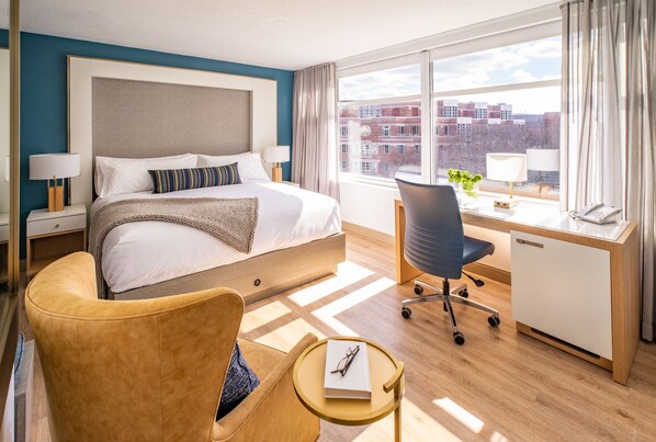 A hotel room with a bed, a desk, a chair, and a view of buildings through large windows.