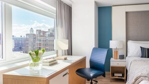 A hotel room with a desk, chair, bed, bedside tables, lamps, and a view of the city.