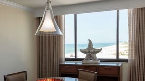 Un espace repas comprenant une table, des chaises, un vase décoratif, une suspension, une sculpture d’étoile de mer et une vue sur la plage grâce à de grandes fenêtres.
