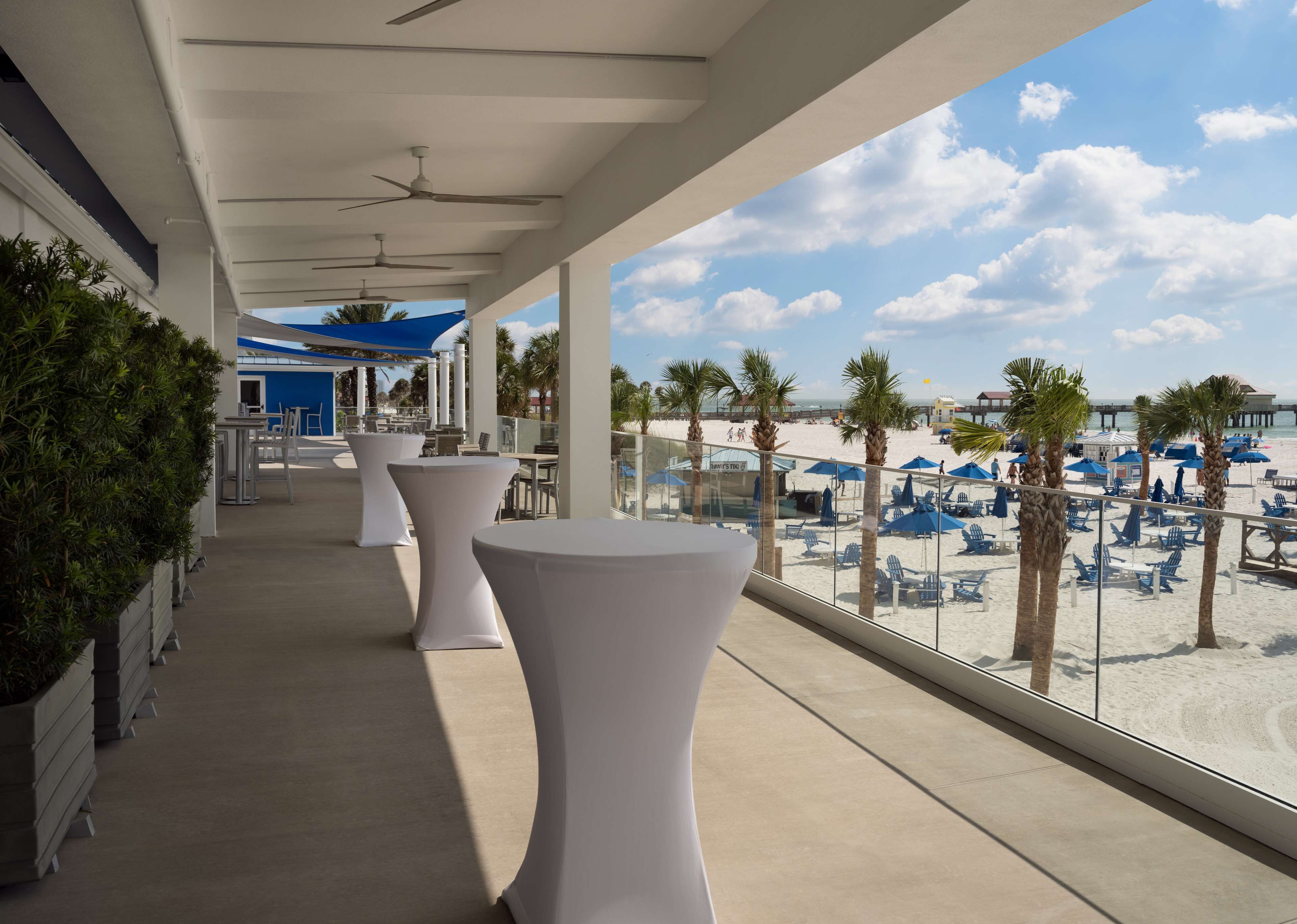 A covered outdoor area with white tables and chairs, overlooking a beach with palm trees and umbrellas.