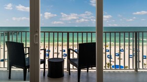 Un balcon avec deux fauteuils en osier et une petite table ronde, donnant sur une plage parsemée de parasols et où se trouvent des gens.