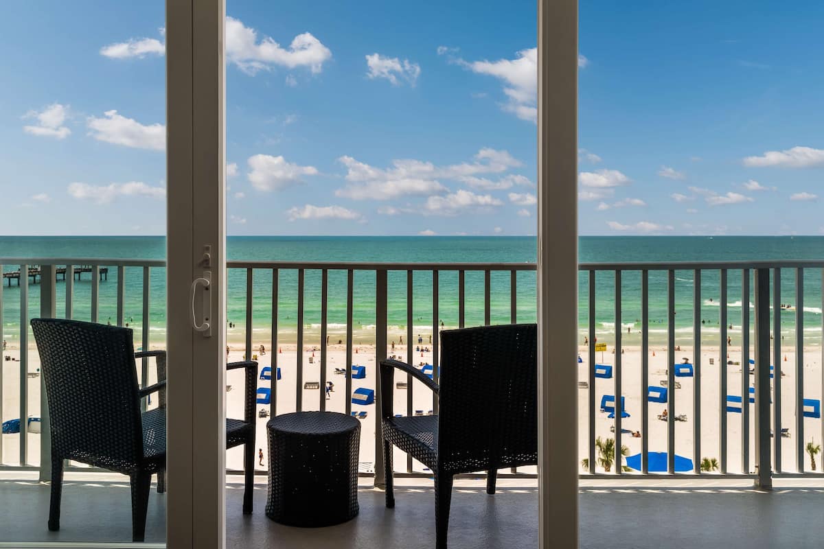 Un balcon avec deux fauteuils en osier et une petite table ronde, donnant sur une plage parsemée de parasols et où se trouvent des gens.