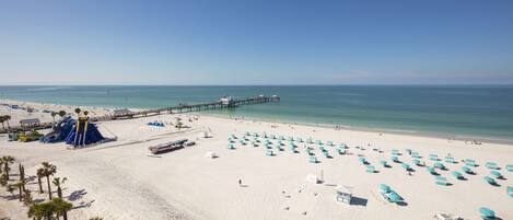 A beach with white sand, clear blue water, and a pier extending into the sea. There are numerous blue umbrellas and lounge chairs arranged on the sand.