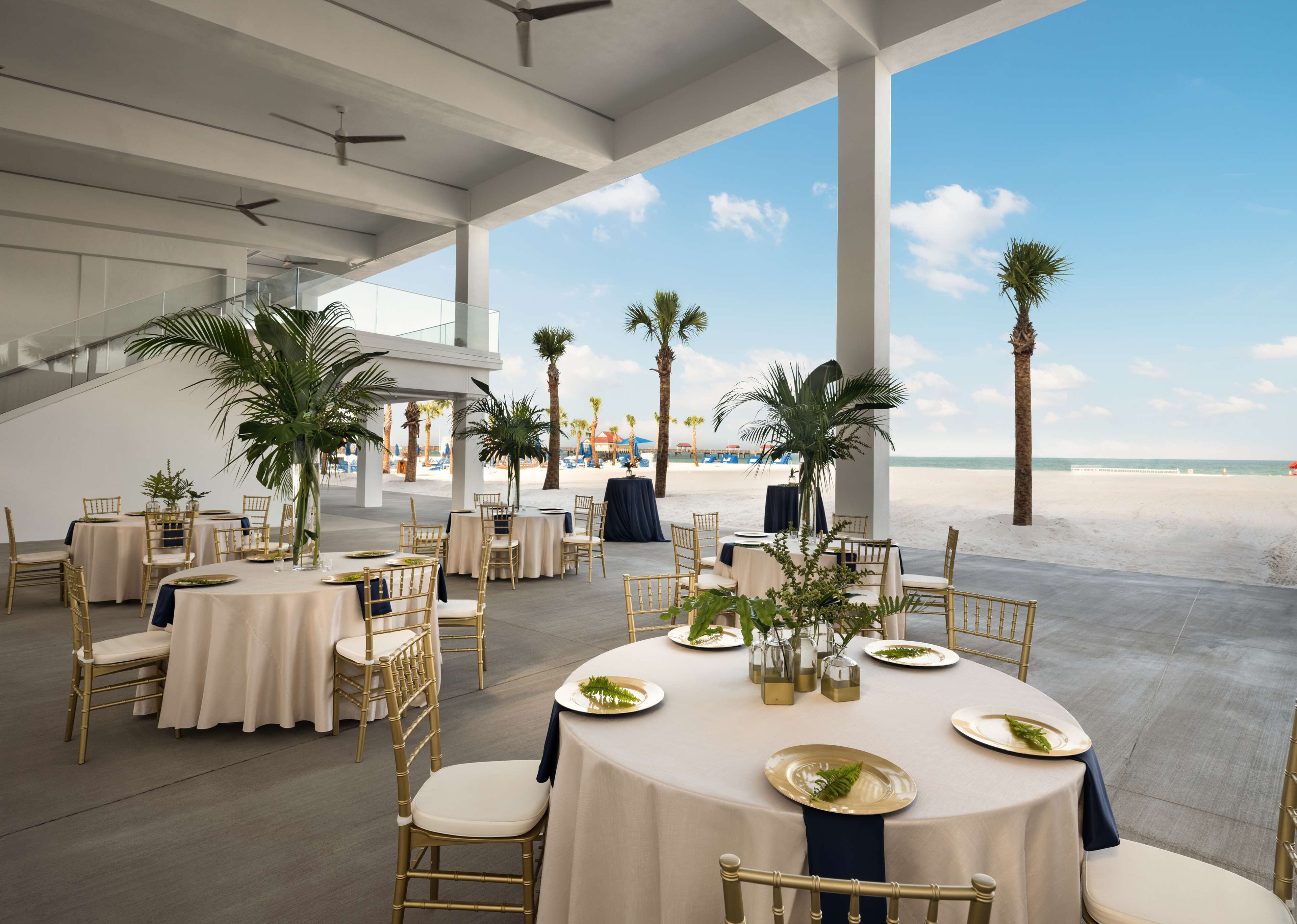 Un área de comedor al aire libre con mesas redondas y sillas doradas, con vista a una playa con palmeras y sombrillas.