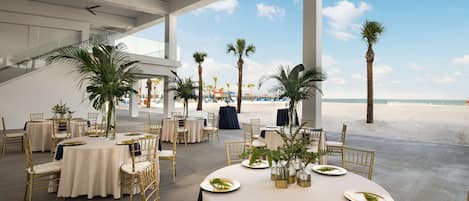 Un área de comedor al aire libre con mesas redondas y sillas doradas, con vista a una playa con palmeras y sombrillas.