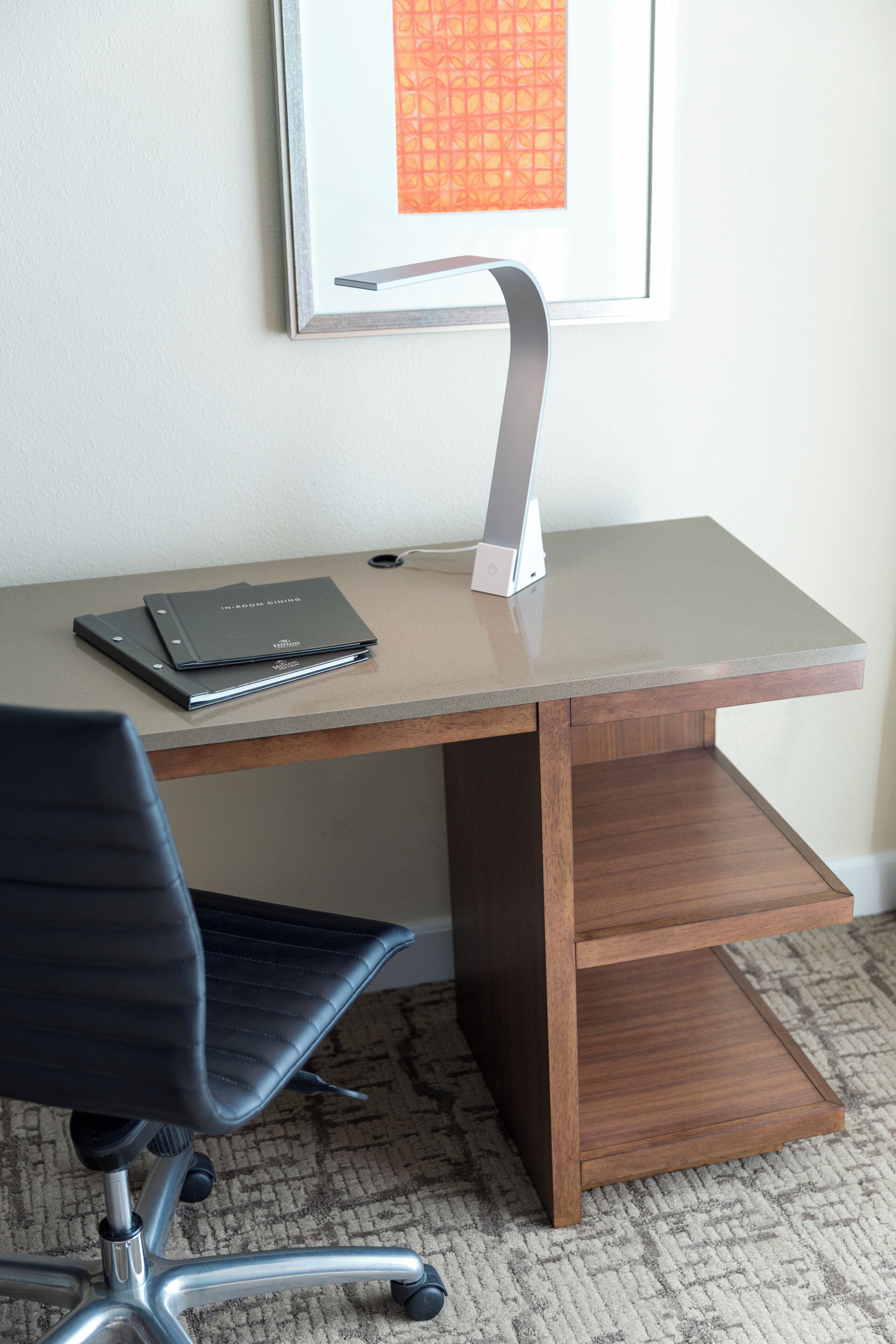A modern desk with a laptop, a notebook, and a pen, a black office chair, and a framed picture on the wall.