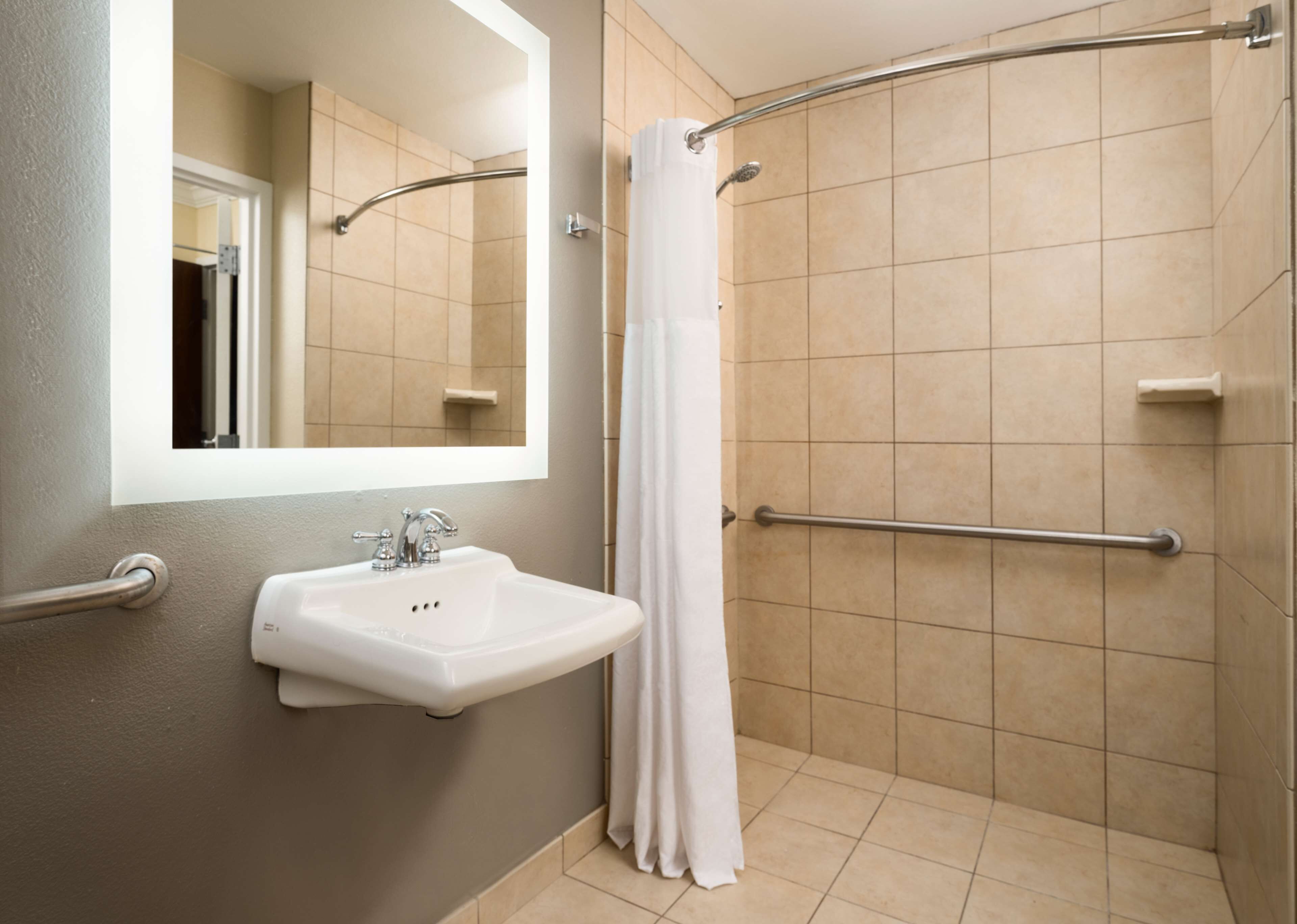 Un baño con lavamanos blanco, un espejo, una ducha con barra de apoyo y paredes con azulejos color crema.