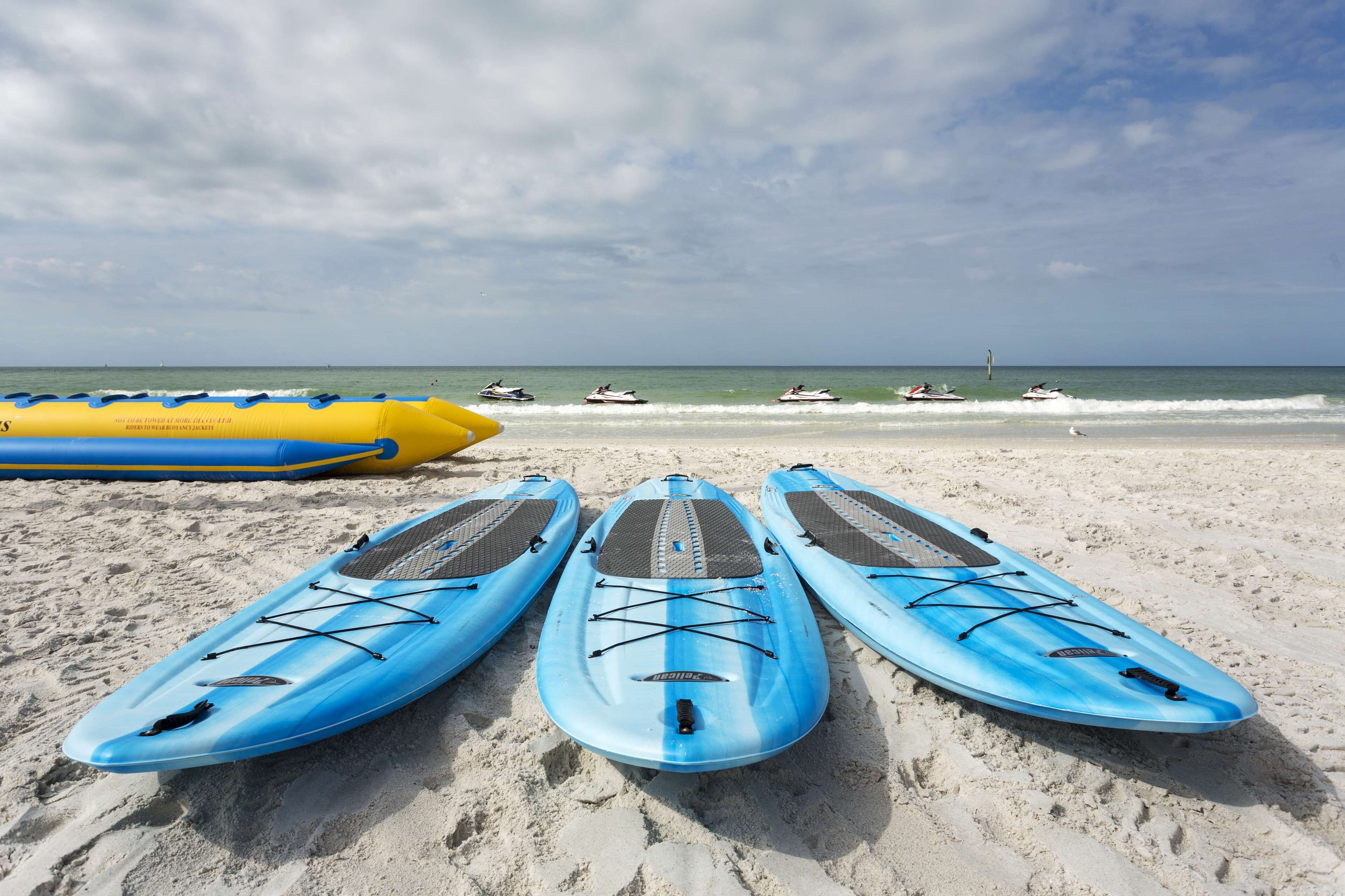 Tres tablas de paddle surf azules sobre una playa arenosa, con vista al océano y motos acuáticas a lo lejos.
