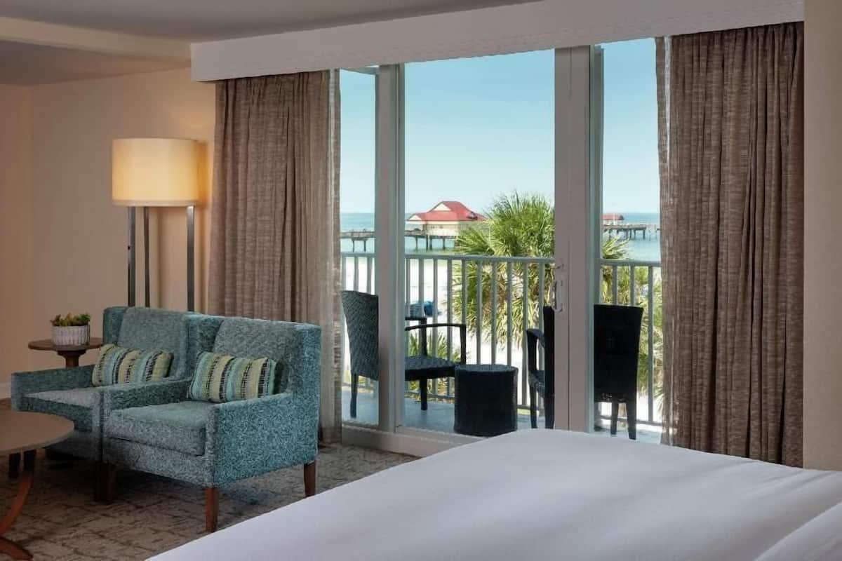 Une chambre d’hôtel équipée d’un lit, d’un canapé, d’une table, d’une lampe et offrant une vue sur l’océan grâce à de grandes fenêtres.