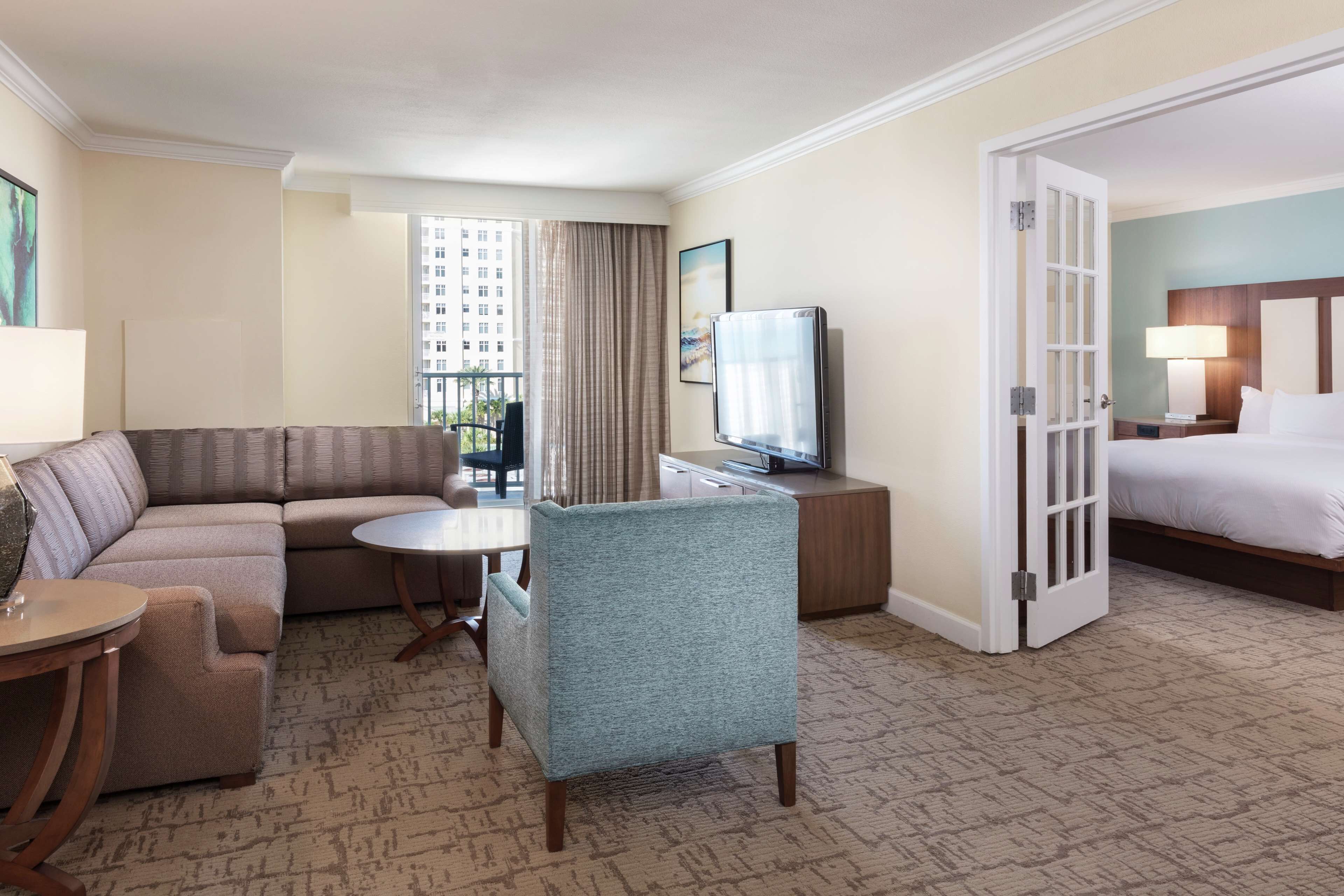 A hotel room with a sofa, a chair, a small table, a TV, a bed, and a door leading to another room.