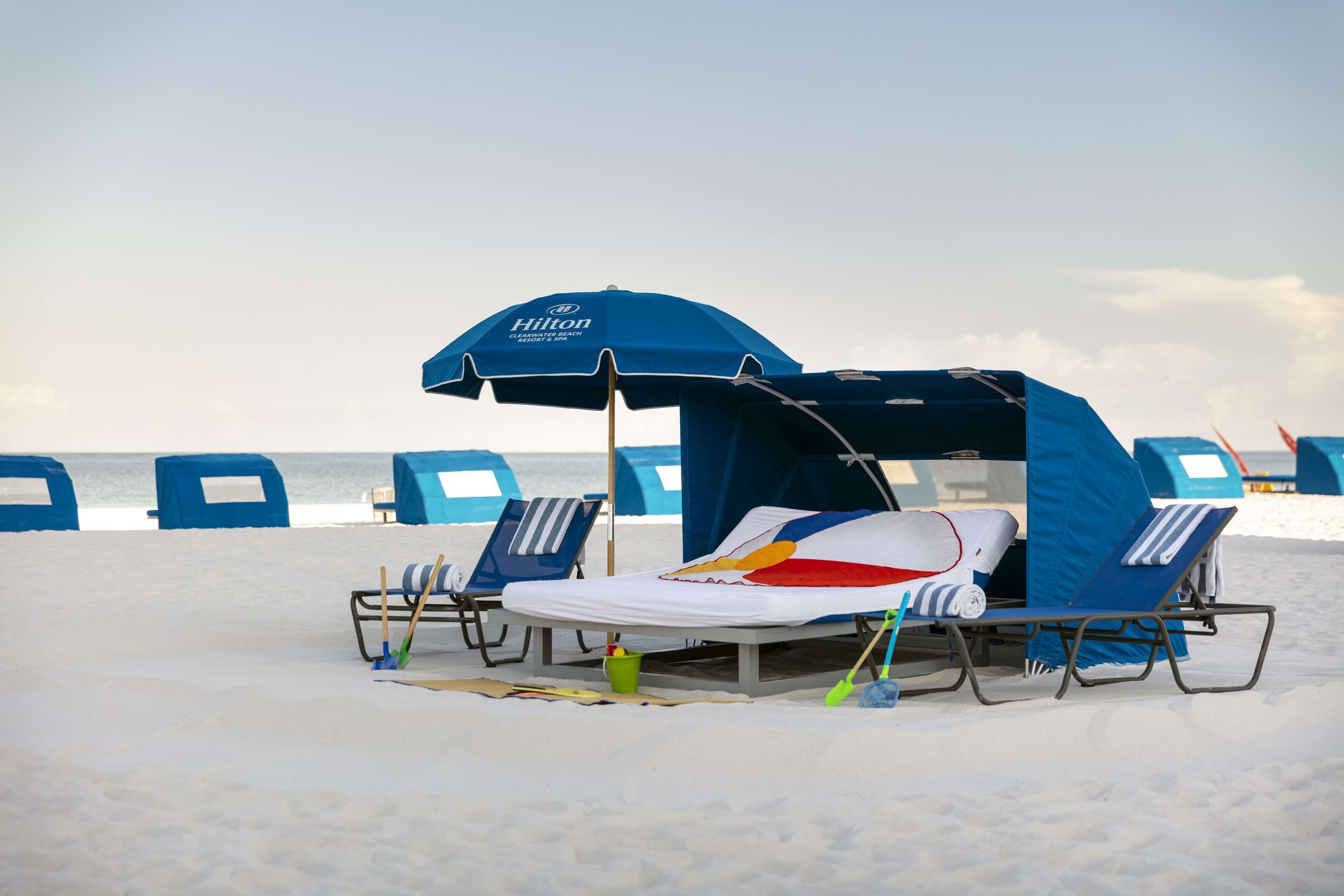 A beachside hotel room with a Hilton canopy, two lounge chairs, and a beach umbrella.
