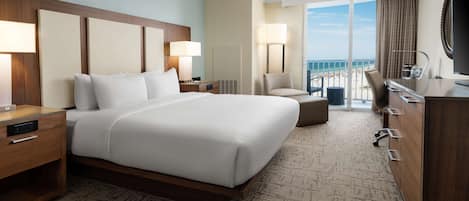 A hotel room with a large bed, a desk with a lamp, a chair, a dresser, and a view of the beach.