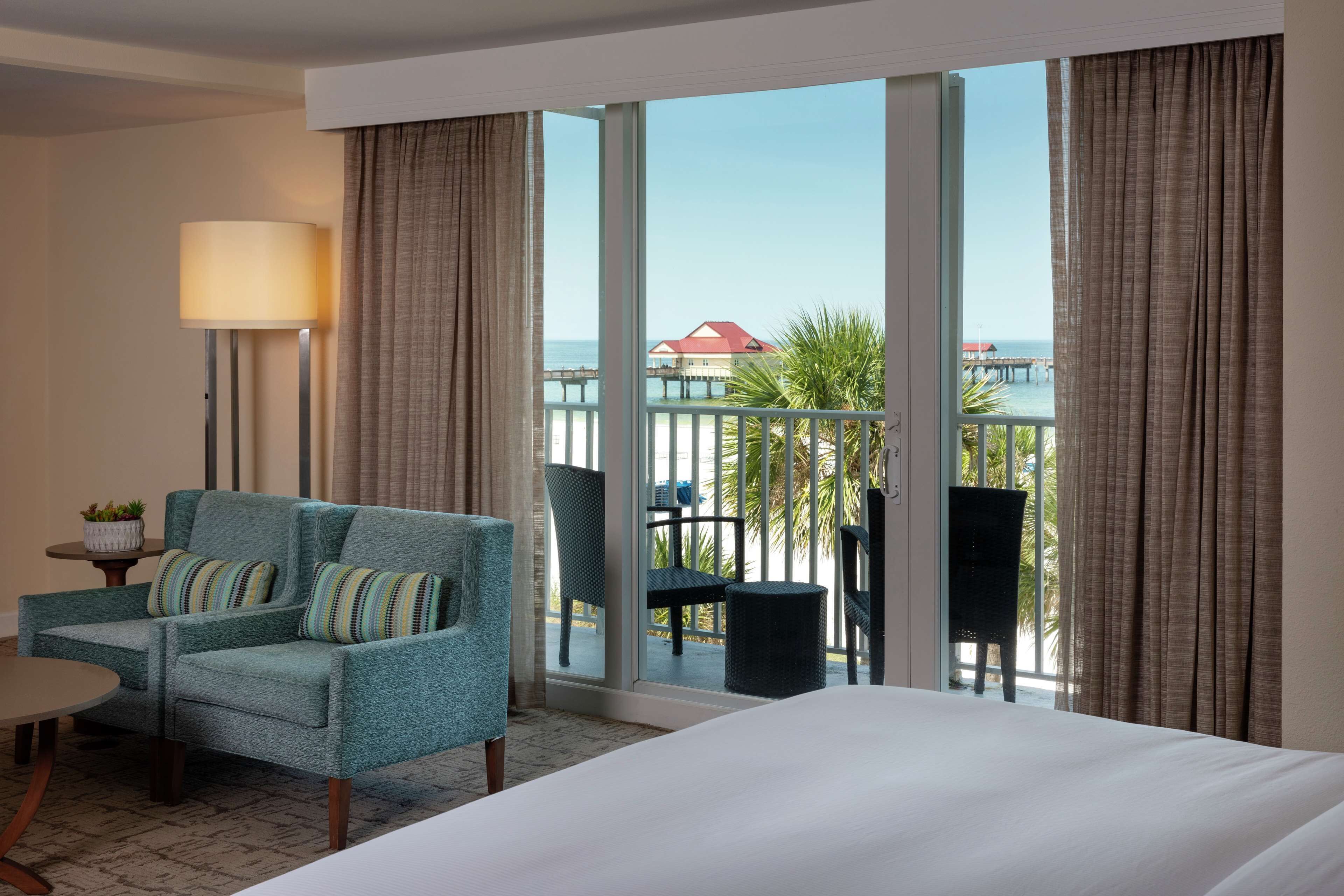 A hotel room with a large bed, a teal armchair with striped cushions, a small table with a lamp, and a view of the ocean through large windows.