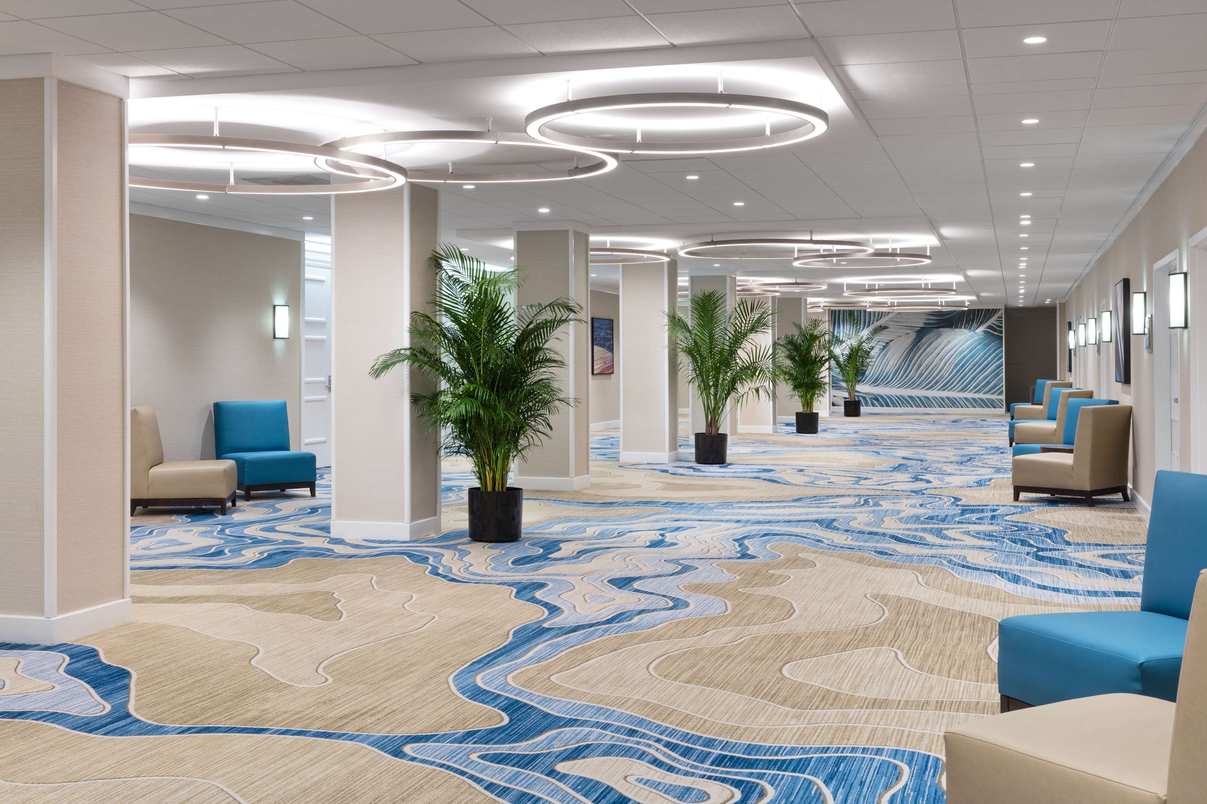 A modern hotel lobby with a patterned carpet, seating areas, and potted plants.