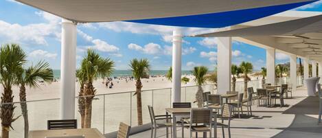 Een terras aan het strand met tafels en stoelen, palmbomen en uitzicht op het strand en de oceaan.