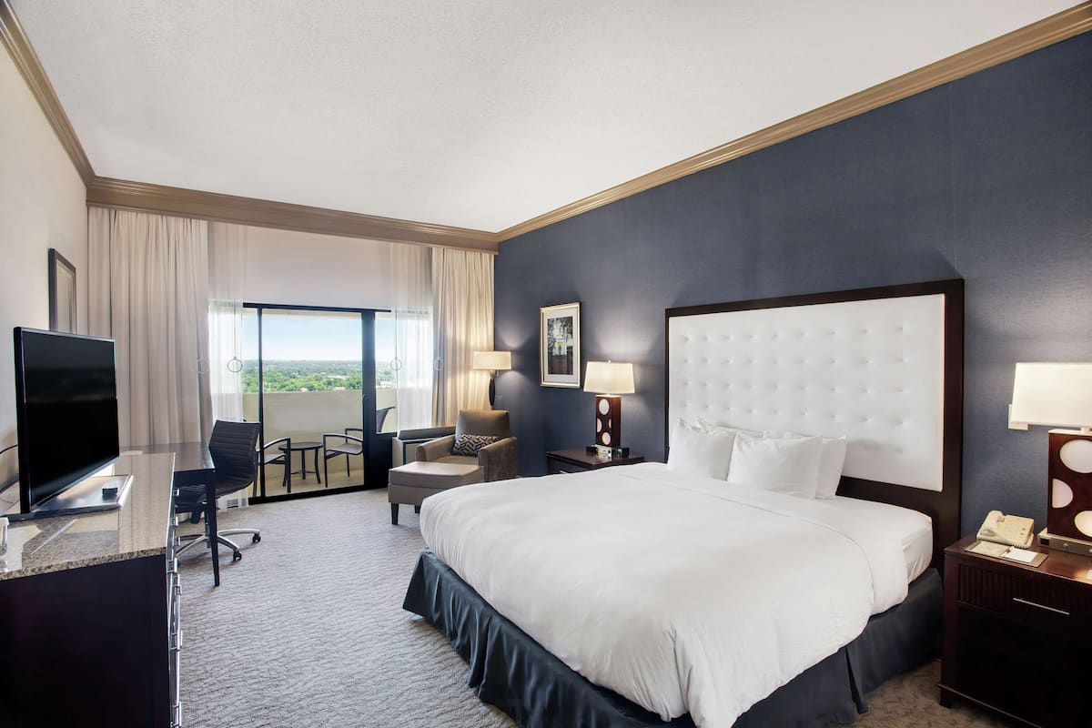 A hotel room with a large bed, a desk with a flat-screen TV, a chair, a small table, and a window with curtains.