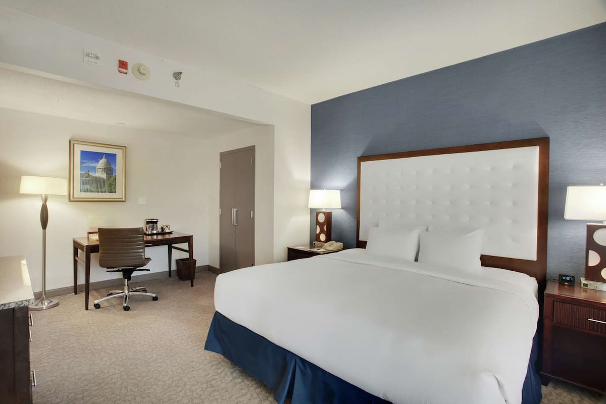 A hotel room with a large bed, a desk, a chair, a lamp, and a clock.