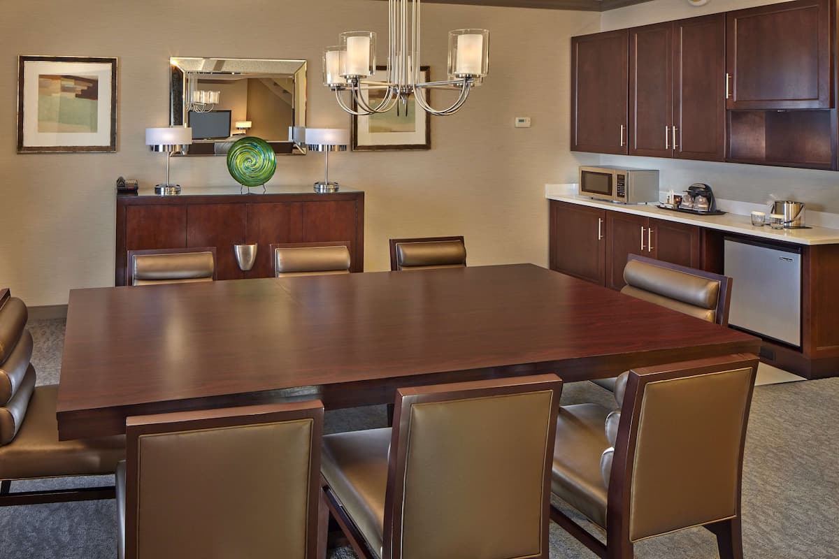 A modern dining area with a large wooden table, leather chairs, a chandelier, and a microwave in the background.