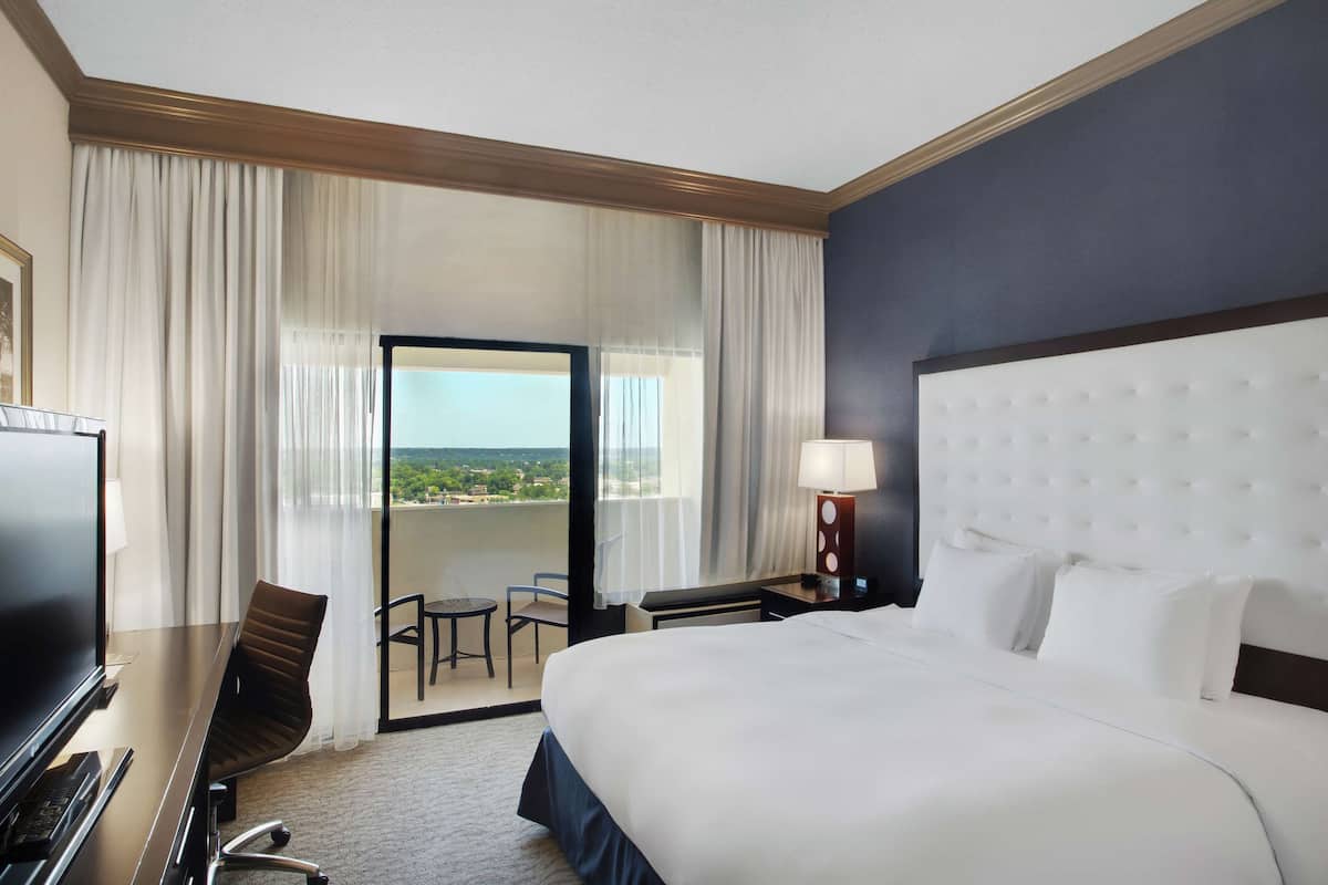 A hotel room with a large bed, a desk with a computer, a chair, a small table, and a view of the outdoors through a sliding door.