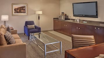 A hotel room with a sofa, armchair, coffee table, dining table, TV, microwave, and a framed picture on the wall.