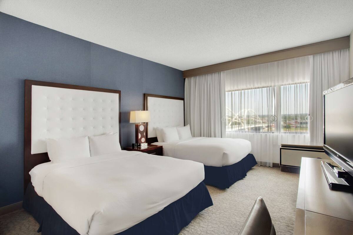 A hotel room with two beds, a desk, a TV, and a window with curtains.
