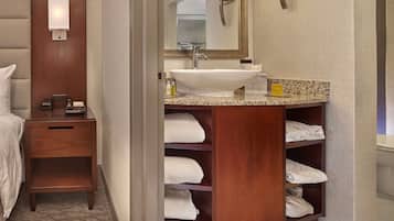 A hotel bathroom with a wooden vanity, a large sink, and a mirror. There are towels stored in a wooden shelf below the sink.