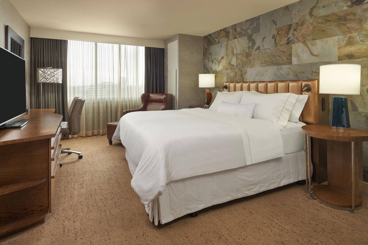 A hotel room with a large bed, a desk, a chair, and a stone wall.