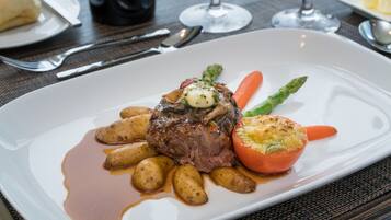 Un plato blanco con un filete cubierto de un champiñón y un tomate pequeño, acompañado de patatas asadas y espárragos.