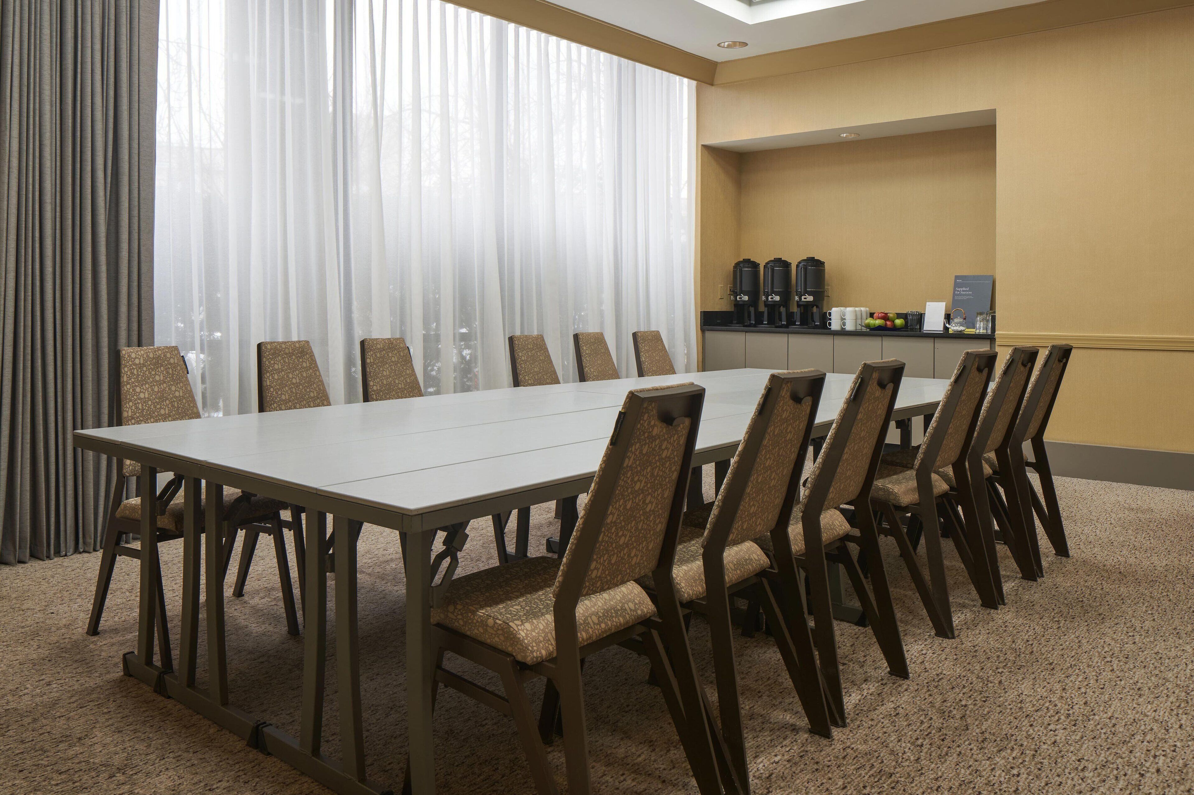 A conference room with a long table and chairs, a coffee station, and a view through sheer curtains.