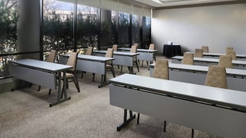 A conference room with multiple tables and chairs, large windows with a view of trees, and a carpeted floor.