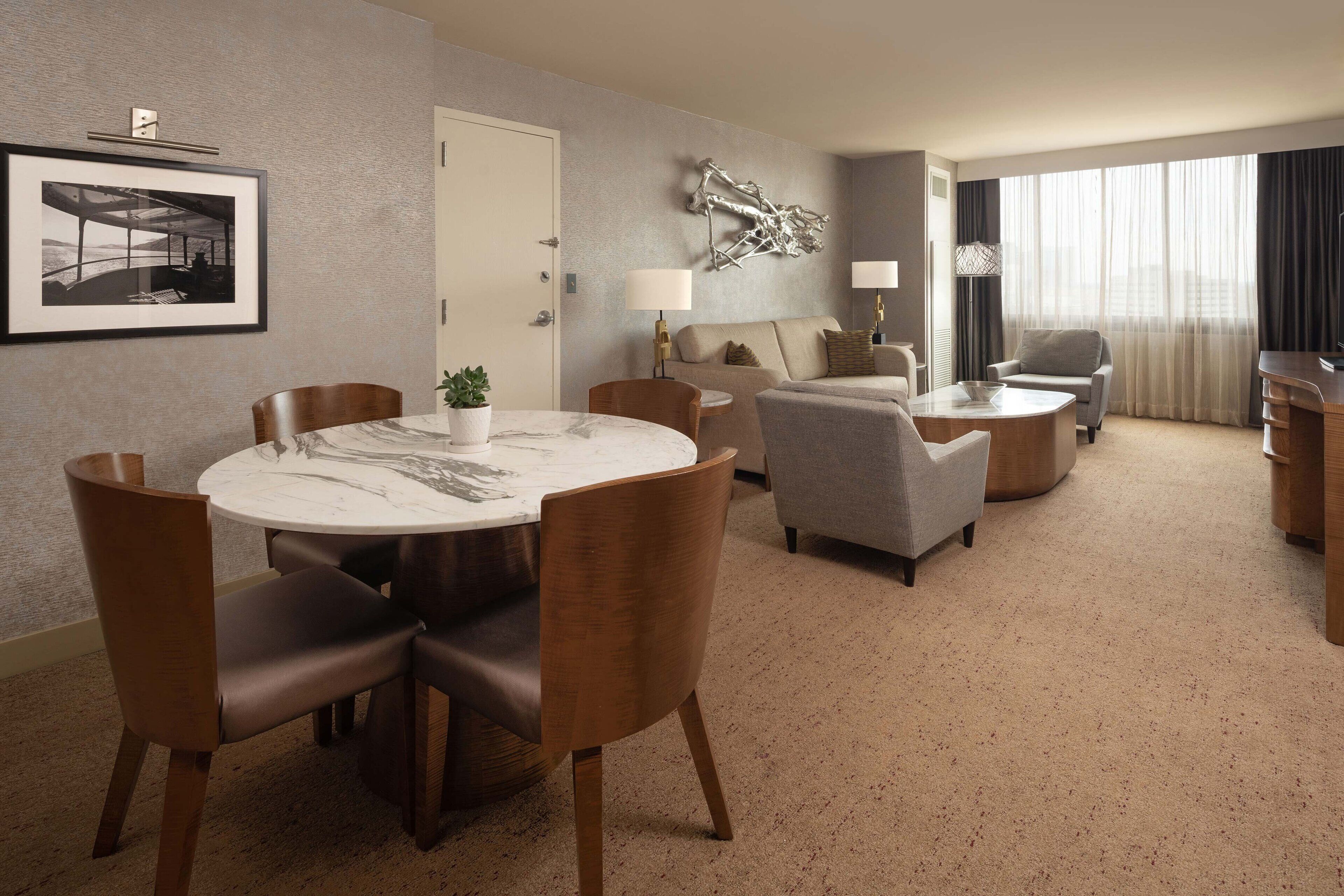 Habitación de hotel con comedor, sofá, sillón, lámpara y un amplio ventanal con cortinas.